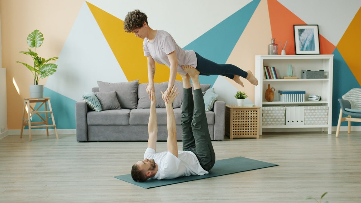 Happy couple is doing pair yoga at home, man is holding woman with legs on floor, girl is stretching arms. Relationship, active people and sports concept.