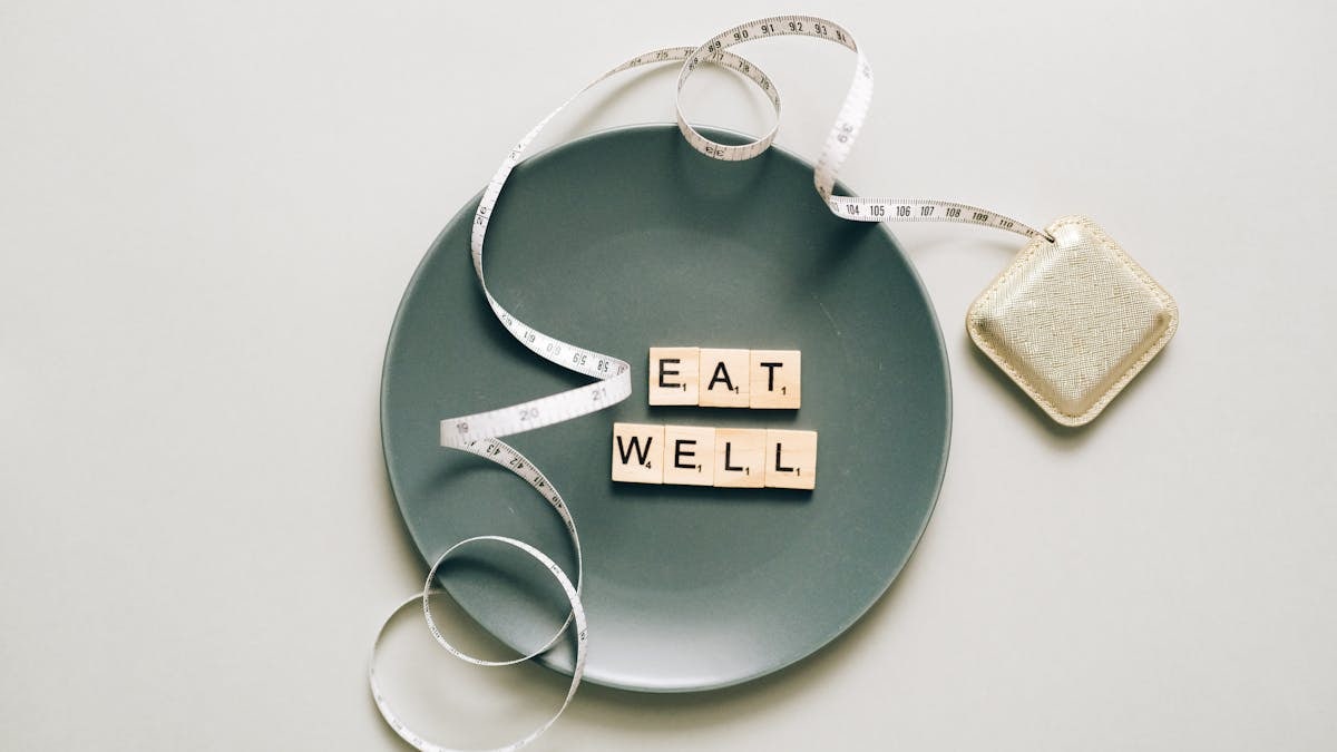 Flat lay of scrabble tiles spelling 'eat well' on a plate with a tape measure, promoting healthy eating.